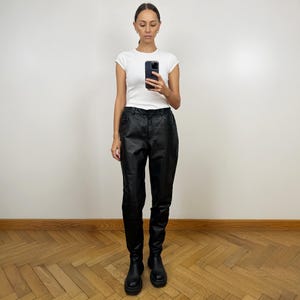 Peut inclure: Une femme portant un t-shirt blanc et un pantalon en cuir noir. Elle est debout sur un sol en bois et regarde son t&eacute;l&eacute;phone.