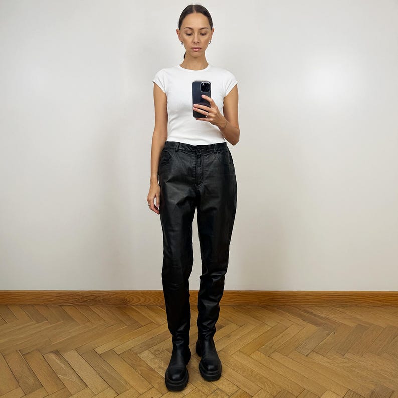 Peut inclure: Une femme portant un t-shirt blanc et un pantalon en cuir noir. Elle est debout sur un sol en bois et regarde son t&eacute;l&eacute;phone.