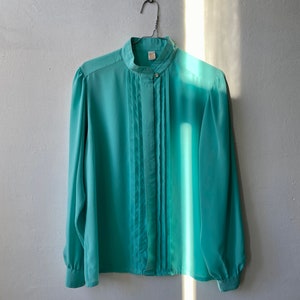 May include: A light teal blouse with a high collar and pleated front. The long-sleeved top has button closures and is hanging on a metal hanger against a white wall. The fabric appears to be lightweight and flowing.