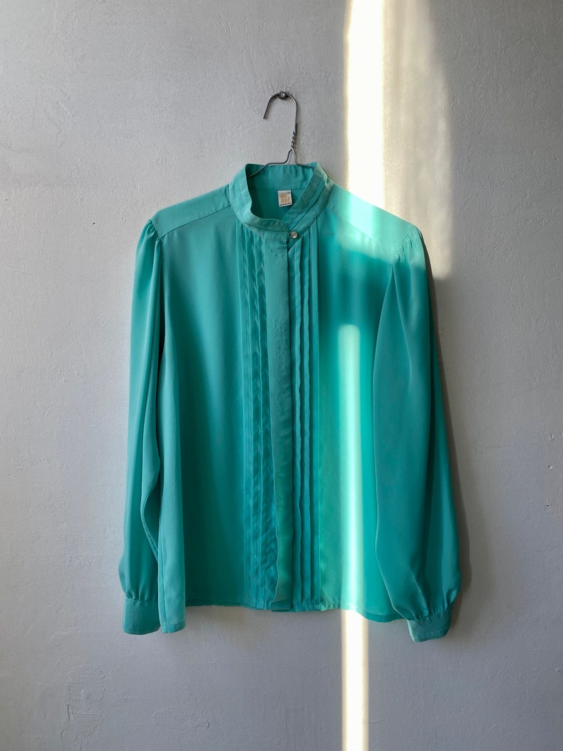 May include: A light teal blouse with a high collar and pleated front. The long-sleeved top has button closures and is hanging on a metal hanger against a white wall. The fabric appears to be lightweight and flowing.