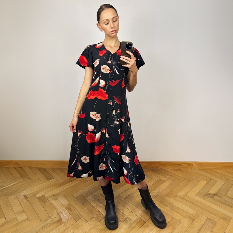 Peut inclure: Une femme portant une robe midi noire &agrave; imprim&eacute; floral rouge et blanc. La robe a un devant boutonn&eacute; et des manches courtes. Elle porte des bottines noires &agrave; la cheville.