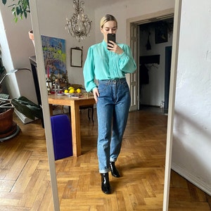 May include: A light turquoise blouse with pleated detailing and long sleeves is paired with high-waisted blue jeans. The outfit is completed with black ankle boots. The image is taken in a room with a mirror.