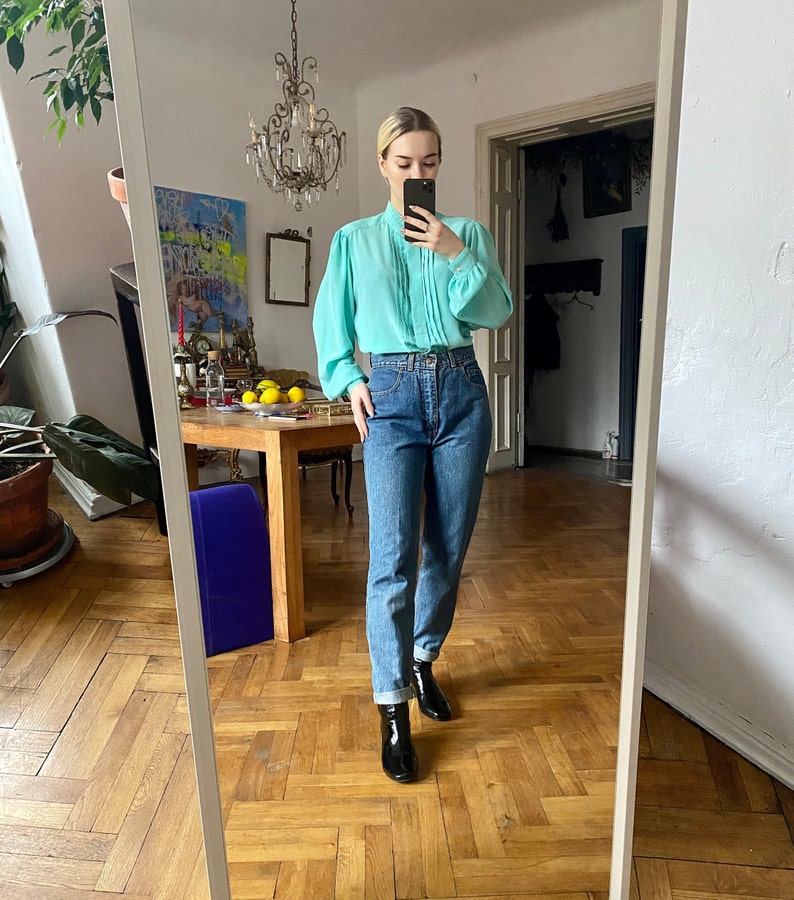 May include: A light turquoise blouse with pleated detailing and long sleeves is paired with high-waisted blue jeans. The outfit is completed with black ankle boots. The image is taken in a room with a mirror.