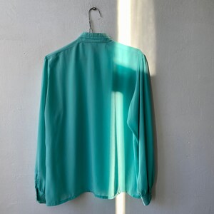 May include: A long-sleeved, light teal blouse hangs on a wire hanger against a white wall. The blouse has a classic collar and buttoned cuffs. The fabric appears lightweight and slightly translucent, with a subtle sheen.