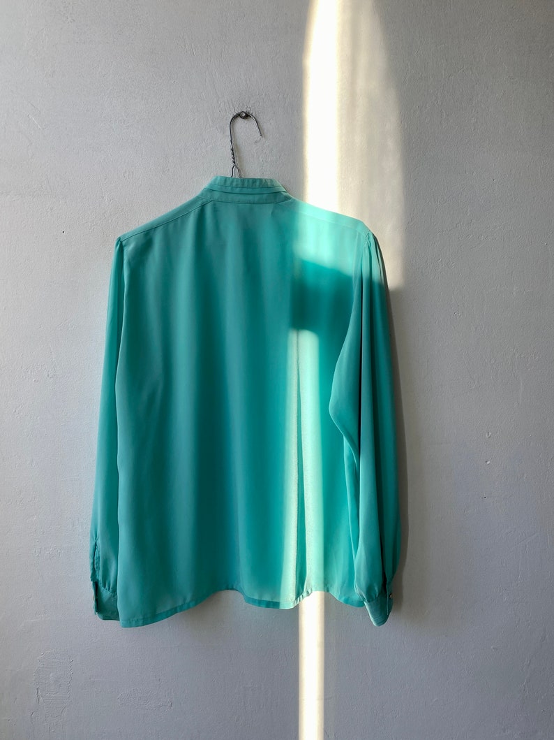 May include: A long-sleeved, light teal blouse hangs on a wire hanger against a white wall. The blouse has a classic collar and buttoned cuffs. The fabric appears lightweight and slightly translucent, with a subtle sheen.