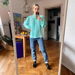 May include: A light turquoise blouse with pleated detailing down the front and long, puffed sleeves. The blouse is paired with blue jeans and black ankle boots. The outfit is reflected in a mirror, showcasing a vintage style.