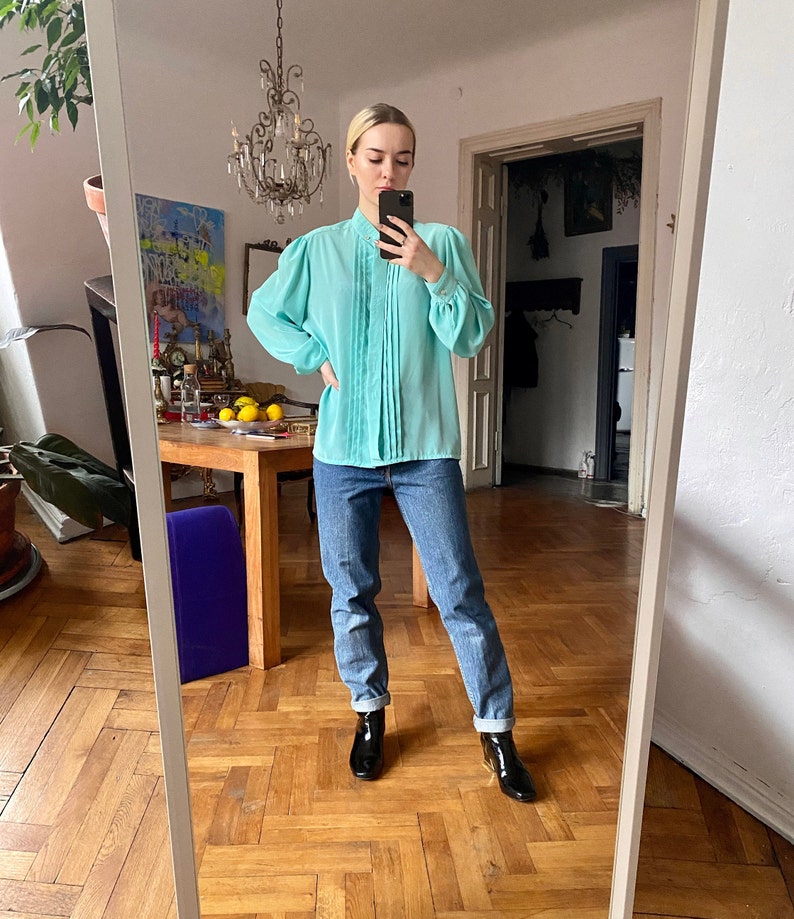 May include: A light turquoise blouse with pleated detailing down the front and long, puffed sleeves. The blouse is paired with blue jeans and black ankle boots. The outfit is reflected in a mirror, showcasing a vintage style.