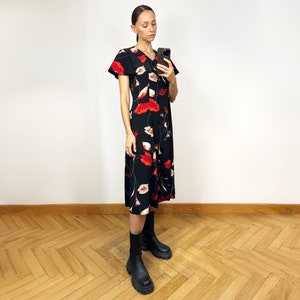 Peut inclure: Une femme portant une robe midi noire &agrave; imprim&eacute; floral rouge et des bottines noires &agrave; la cheville. Elle tient un smartphone noir dans sa main droite.