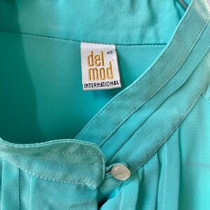 May include: A close-up of a light turquoise blouse with a mandarin collar and a single button closure. The label reads "del mod INTERNATIONAL" in gold lettering. The blouse is hanging on a metal hanger.