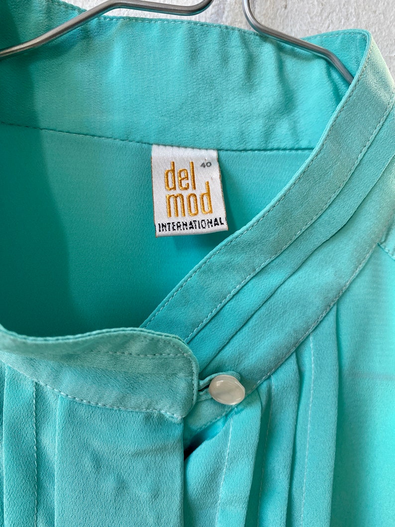 May include: A close-up of a light turquoise blouse with a mandarin collar and a single button closure. The label reads "del mod INTERNATIONAL" in gold lettering. The blouse is hanging on a metal hanger.