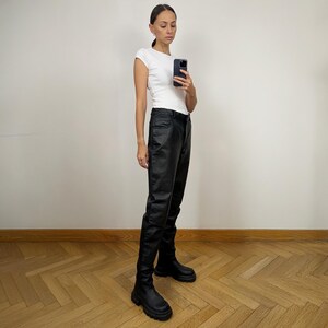 Peut inclure: Une femme portant un t-shirt blanc et un pantalon en cuir noir. Elle porte des bottines noires &agrave; la cheville et tient un smartphone noir.
