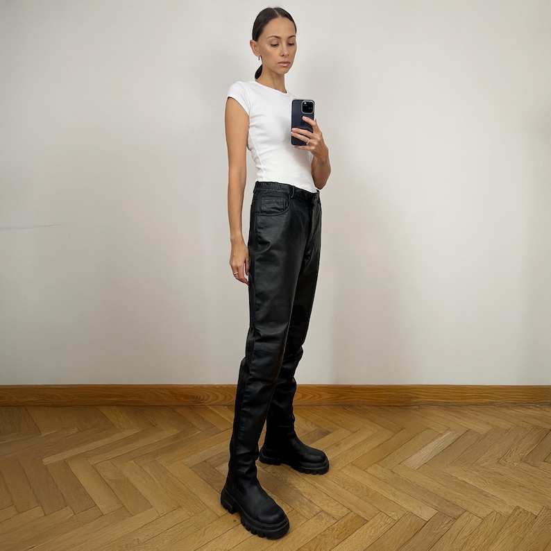 Peut inclure: Une femme portant un t-shirt blanc et un pantalon en cuir noir. Elle porte des bottines noires &agrave; la cheville et tient un smartphone noir.