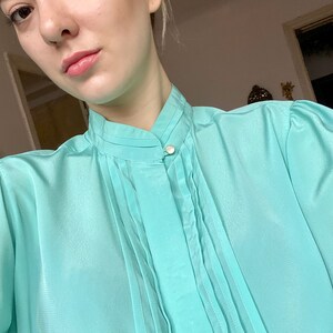 May include: A light turquoise blouse with a high collar and pleated front detail. The blouse has long sleeves and a single pearl button closure at the neck. The fabric appears to be a lightweight, semi-sheer material.