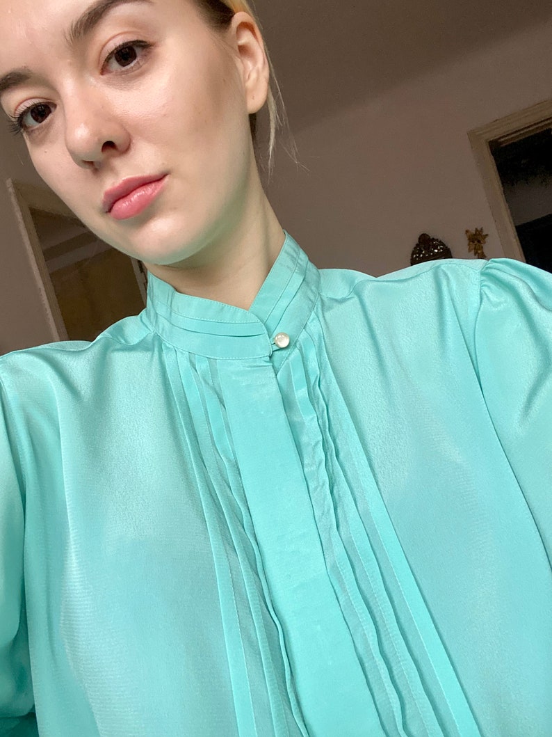 May include: A light turquoise blouse with a high collar and pleated front detail. The blouse has long sleeves and a single pearl button closure at the neck. The fabric appears to be a lightweight, semi-sheer material.
