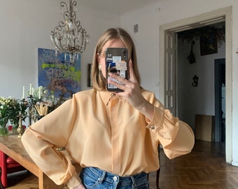 May include: A light peach-colored button-down blouse with long sleeves and gold-toned button details. The blouse is paired with blue denim jeans. The sleeves have a slight puff at the shoulders and gather at the cuffs.