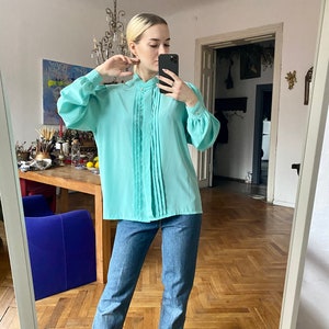 May include: A light turquoise blouse with pleated detailing down the front and long, voluminous sleeves. The blouse has a high neckline and is paired with blue jeans. The outfit is completed with black ankle boots.