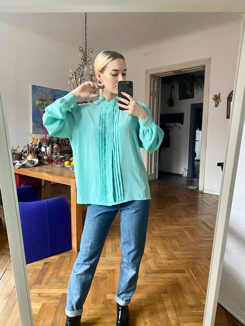 May include: A light turquoise blouse with pleated detailing down the front and long, voluminous sleeves. The blouse has a high neckline and is paired with blue jeans. The outfit is completed with black ankle boots.