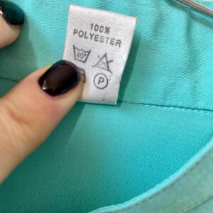 May include: Close-up of a label on a teal garment. The label reads "100% POLYESTER" and includes washing instructions: a wash symbol with 40 degrees, a do not bleach symbol, and a dry cleaning symbol.