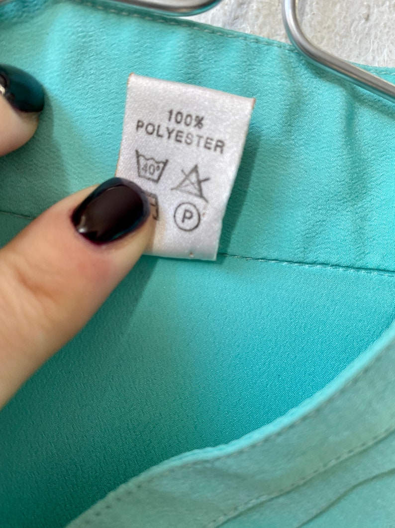 May include: Close-up of a label on a teal garment. The label reads "100% POLYESTER" and includes washing instructions: a wash symbol with 40 degrees, a do not bleach symbol, and a dry cleaning symbol.