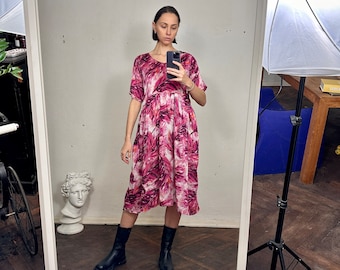 May include: A knee-length dress with a pink and white leaf pattern. The dress has short sleeves and a gathered waist. The person in the image is wearing black boots. The dress is reflected in a large mirror.