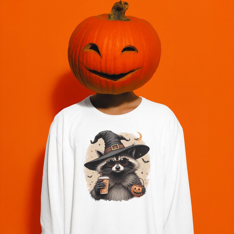 May include: A white long-sleeve shirt featuring a graphic of a raccoon wearing a black witch hat and sunglasses. The raccoon holds a coffee cup and a small jack-o'-lantern. The background of the graphic includes small bats and a crescent moon.