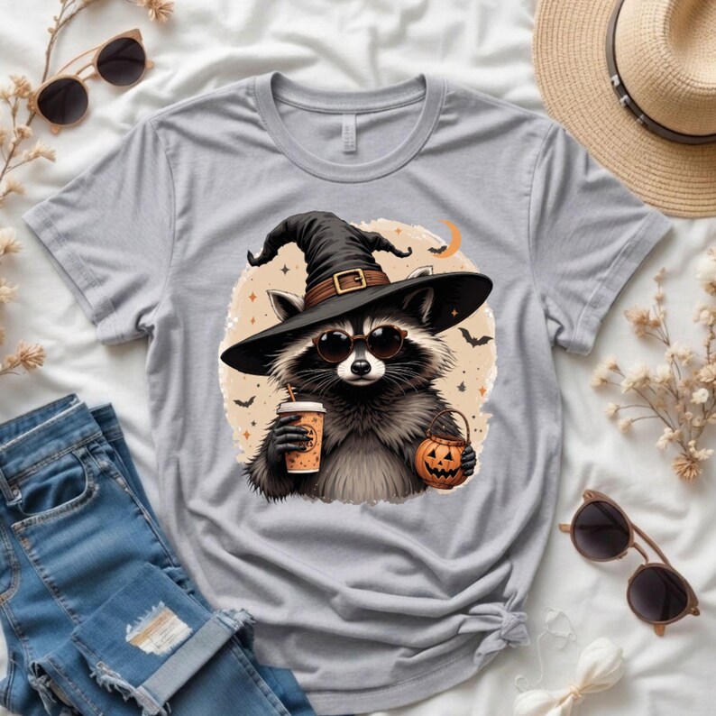 May include: A heather gray t-shirt featuring a graphic of a raccoon wearing a black witch hat and round sunglasses. The raccoon holds a coffee cup and a small orange jack-o'-lantern bucket. The design includes small bats and a crescent moon.