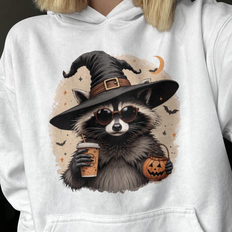 May include: A white hoodie featuring a graphic of a raccoon wearing a black witch hat and round sunglasses. The raccoon holds a tan coffee cup in one hand and a small orange jack-o'-lantern bucket in the other, set against a beige circular background.
