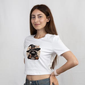 May include: A white short-sleeved t-shirt featuring a graphic print of a raccoon wearing a black witch hat and sunglasses, holding a coffee cup and a small orange pumpkin. The design includes a crescent moon and small bat silhouettes in the background.