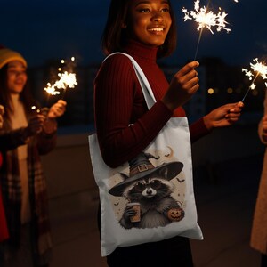 May include: A white tote bag featuring a graphic illustration of a raccoon wearing a witch hat and sunglasses, holding a coffee cup and a jack-o'-lantern bucket, surrounded by small bats and a crescent moon against a light beige background.