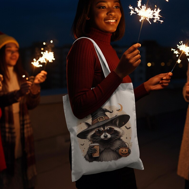 May include: A white tote bag featuring a graphic illustration of a raccoon wearing a witch hat and sunglasses, holding a coffee cup and a jack-o'-lantern bucket, surrounded by small bats and a crescent moon against a light beige background.