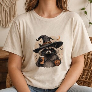 May include: A cream-colored t-shirt featuring a graphic of a raccoon wearing a black witch hat and round sunglasses. The raccoon holds a coffee cup and a small jack-o'-lantern bucket, surrounded by small stars, bats, and a crescent moon.