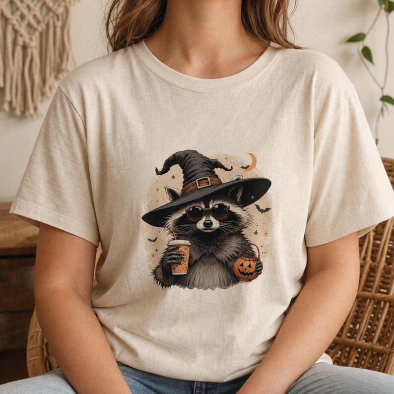 May include: A cream-colored t-shirt featuring a graphic of a raccoon wearing a black witch hat and round sunglasses. The raccoon holds a coffee cup and a small jack-o'-lantern bucket, surrounded by small stars, bats, and a crescent moon.