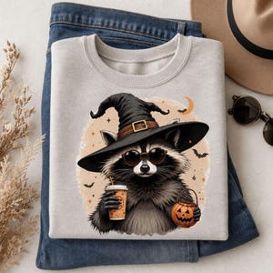 May include: A light gray crewneck sweatshirt featuring a graphic of a raccoon wearing a black witch hat and round sunglasses. The raccoon holds a coffee cup in one hand and a small orange jack-o'-lantern bucket in the other, set against a moon.