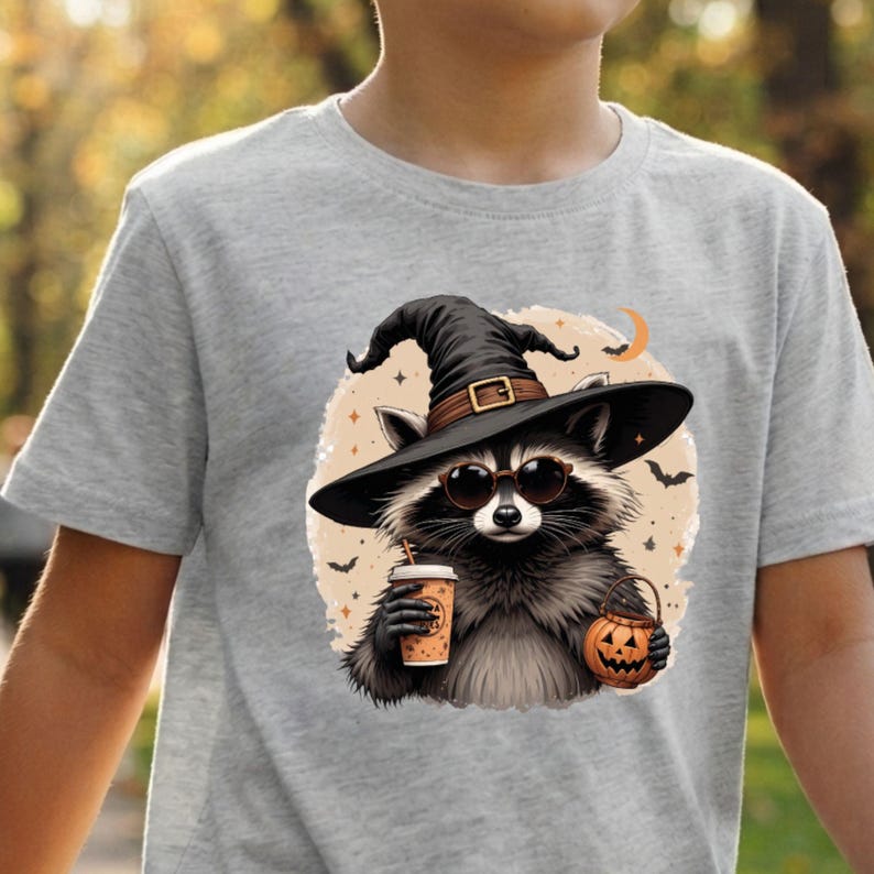 May include: A heather gray t-shirt featuring a graphic of a raccoon wearing a black witch hat and round sunglasses. The raccoon holds a tan coffee cup and a small orange jack-o'-lantern bucket. The background includes bats and a crescent moon.