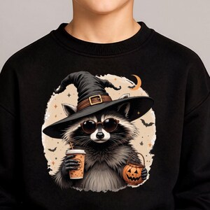 May include: A black sweatshirt featuring a graphic of a raccoon wearing a black witch hat and round sunglasses. The raccoon holds a tan coffee cup in one hand and a small orange jack-o'-lantern bucket in the other, set against a beige circle.