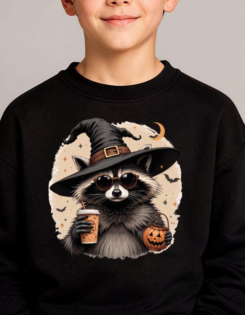 May include: A black sweatshirt featuring a graphic of a raccoon wearing a black witch hat and round sunglasses. The raccoon holds a tan coffee cup in one hand and a small orange jack-o'-lantern bucket in the other, set against a beige circle.