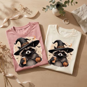 May include: Two t-shirts, one dusty pink and one cream, each featuring a graphic of a raccoon wearing a black witch hat and sunglasses. The raccoon holds a coffee cup and a small jack-o'-lantern bucket against a circular background with bats.