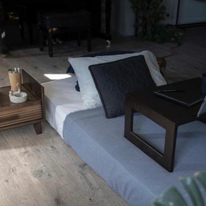 Può includere: Un tavolino da salotto in legno scuro con un cassetto e un libro intitolato "Fashion" sopra. Una coperta blu con un cuscino nero è sul pavimento accanto al tavolo. Un laptop e una tazza di caffè sono su un tavolino da portata in legno nero.