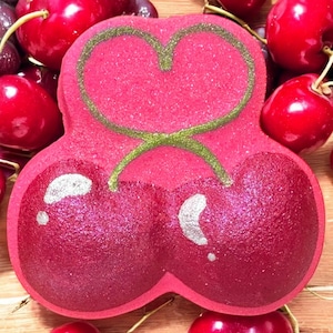 May include: A vibrant, cherry-shaped bath bomb with a heart-shaped design. The bath bomb is a deep red color with a glittery finish, and a green stem and heart outline. Fresh cherries surround the bath bomb.
