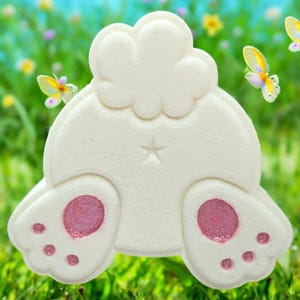 May include: A white, bunny-shaped bath bomb with pink paw pads and a star detail. The bath bomb is set against a blurred background of green grass and colorful butterflies, suggesting a spring or Easter theme.