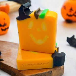May include: Two orange pumpkin-shaped soaps with a black witch hat and green ribbon on top. The soap on top has a smiling jack-o-lantern face. The bottom soap has the word "Sudncraft" printed on it.