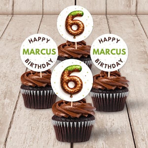 Può includere: Cinque cupcake al cioccolato con glassa al cioccolato e decorazioni. Le decorazioni recitano "Happy Marcus Birthday" in testo verde. Ogni cupcake ha un topper a forma di palloncino con il numero sei. I cupcake sono su una superficie di legno chiaro.