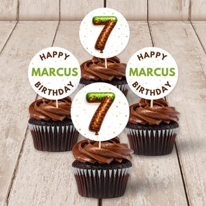 Può includere: Quattro cupcake al cioccolato con glassa al cioccolato e pirottini bianchi. Ogni cupcake ha un topper rotondo con la scritta "HAPPY MARCUS BIRTHDAY" in verde e un numero 7 marrone. I cupcake sono su una superficie di legno chiaro.