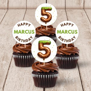 Può includere: Cinque cupcake al cioccolato con glassa al cioccolato e decorazioni. Le decorazioni sono cerchi bianchi con la scritta "Happy Marcus Birthday" in verde e marrone. Ogni cupcake ha un numero 5.
