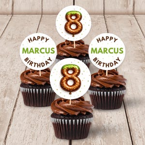 Può includere: Quattro cupcake al cioccolato con glassa al cioccolato e decorazioni. Le decorazioni sono cerchi bianchi con la scritta "HAPPY MARCUS BIRTHDAY" in verde e marrone. Due decorazioni hanno un numero 8 marrone con la parte superiore verde.