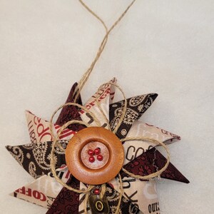 May include: A brown and white fabric star ornament with a brown wooden button and red beads in the center. The star is tied with twine and has the words "Coffee" and "Our" printed on it.