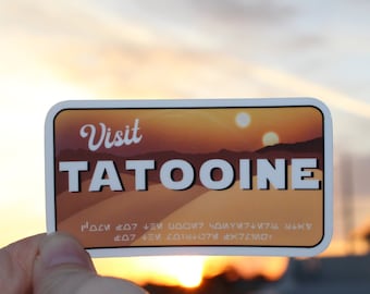 Puede incluir: Una pegatina blanca con una escena de puesta de sol y el texto "Visit Tatooine" en una fuente retro. La pegatina también incluye el texto "This is the way" en Aurebesh, el idioma del universo de Star Wars.