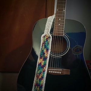 May include: Acoustic guitar with a white and rainbow crochet guitar strap. The strap features a colorful pattern of red, yellow, green, and blue. The guitar is black with a wooden soundhole and bridge.