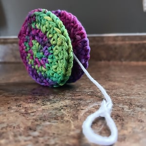 Puede incluir: Un juguete de yoyo de crochet con un dise&ntilde;o multicolor de morado, verde y rosa. El yoyo est&aacute; unido a una cuerda blanca.