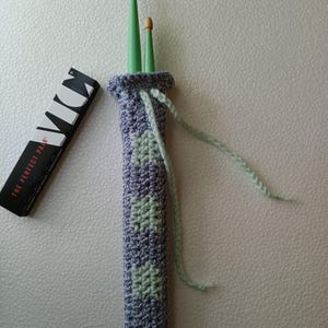 Puede incluir: Un soporte de baquetas de ganchillo en tonos morados y verdes, con un patrón de cuadros. Se ven dos baquetas verdes con puntas de madera. Una caja en blanco y negro con el texto "THE PERFECT PAIR" está a la izquierda.